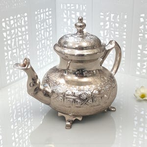 Peut inclure: Une théière argentée avec des motifs floraux et géométriques complexes. La théière a un bec courbé, un corps arrondi, une anse et un couvercle avec un bouton décoratif. Elle repose sur trois petits pieds.