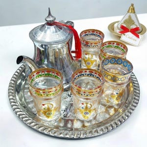 May include: A silver tea set with a teapot, six glasses, and a serving tray. The glasses have colorful patterns. A small gift box with a red ribbon is also included. The set is designed for serving tea.