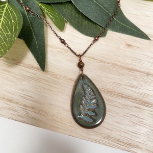 May include: A copper-toned necklace featuring a teardrop pendant with a fern design. The pendant is a muted green with a light blue fern. The necklace chain has small copper beads and a larger bead above the pendant.