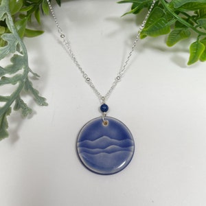 May include: A silver chain necklace with a round, blue pendant. The pendant features a mountain range design. A small, dark blue bead is between the pendant and the chain. Green foliage is in the background.