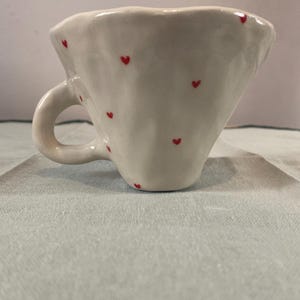 May include: White ceramic coffee cup with a ruffled rim and a handle. The cup is decorated with small red hearts.