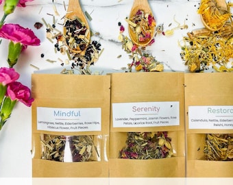 May include: Three brown paper pouches of herbal tea labeled Mindful, Serenity, and Restorative, each with a clear window showing dried tea blends. Three wooden spoons hold loose tea leaves above the pouches, accompanied by bright pink carnations.