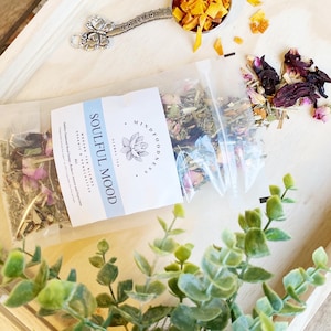 May include: A white pouch with a label that reads "SOULFUL MOOD" and "MINDFOOD" with a floral design. The pouch contains a blend of dried herbs and flowers. The pouch is sitting on a wooden tray with a small spoon filled with dried orange peel.