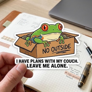 May include: A sticker depicting a cartoon frog with red eyes, peeking out of a cardboard box. The box reads "NO OUTSIDE." The sticker also says, "I HAVE PLANS WITH MY COUCH. LEAVE ME ALONE."