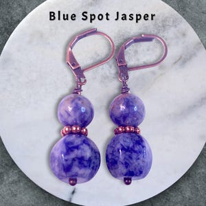 May include: A pair of earrings featuring blue spot jasper beads. The earrings have a lever-back closure and are made with copper-colored metal. The earrings have a two-bead design, with a smaller bead above a larger bead. The text "Blue Spot Jasper" is at the top.