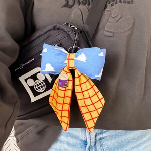 Peut inclure: Un porte-cl&eacute;s n&oelig;ud en tissu attach&eacute; &agrave; un sac bandouli&egrave;re gris fonc&eacute;. Le n&oelig;ud pr&eacute;sente un tissu bleu clair avec des nuages blancs et un tissu jaune avec un motif &agrave; grille rouge. Un petit baume &agrave; l&egrave;vres cylindrique est gliss&eacute; dans une poche du pan jaune.