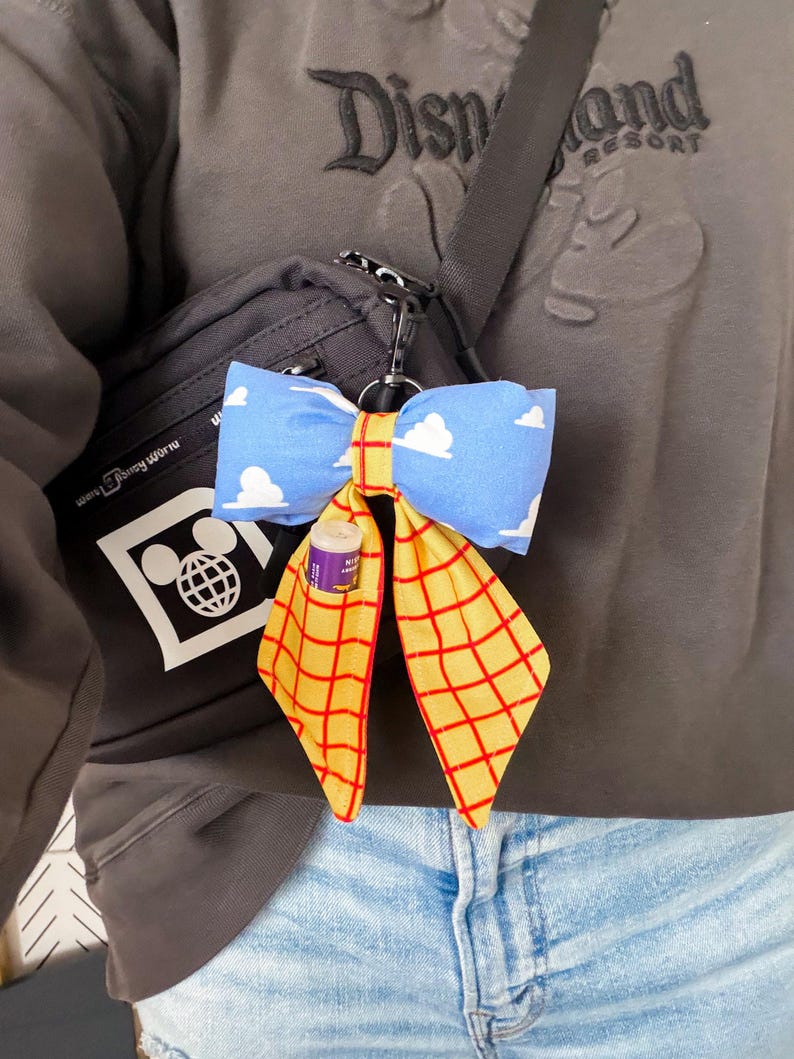 Peut inclure: Un porte-cl&eacute;s n&oelig;ud en tissu attach&eacute; &agrave; un sac bandouli&egrave;re gris fonc&eacute;. Le n&oelig;ud pr&eacute;sente un tissu bleu clair avec des nuages blancs et un tissu jaune avec un motif &agrave; grille rouge. Un petit baume &agrave; l&egrave;vres cylindrique est gliss&eacute; dans une poche du pan jaune.