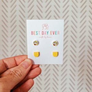 May include: A pair of stud earrings and two yellow bar earrings on a white card. The stud earrings feature a cartoon character design. The card reads "BEST DAY EVER" in colorful text.