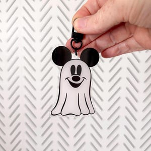May include: A clear acrylic keychain featuring a white ghost character with black mouse ears, black eyes, a black nose, and a wide black smiling mouth. The ghost is attached to a black metal lobster clasp ring for easy attachment to items.