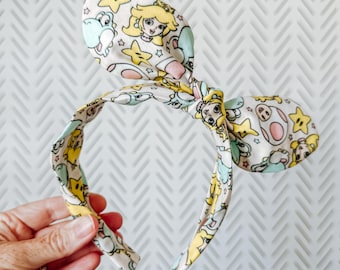 May include: A fabric headband with a bow, featuring a colorful pattern of cartoon characters, including Princess Peach, Yoshi, and stars. The headband is held by a hand against a white and gray chevron background.