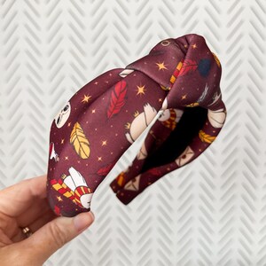 May include: A burgundy headband with a knotted design. The headband features a repeating pattern of white owls, yellow feathers, and gold stars on a dark burgundy background.