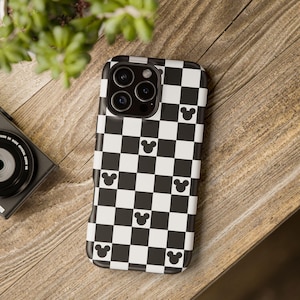 May include: A black and white checkered phone case with Mickey Mouse silhouettes. The case has a glossy finish and a camera cutout. The phone case is on a wooden surface.