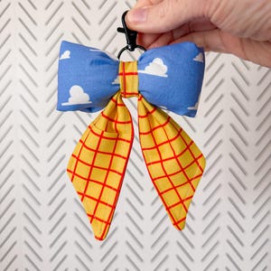 Peut inclure: Un accessoire en forme de n&oelig;ud en tissu avec un motif de nuages blancs sur fond bleu pour la partie sup&eacute;rieure et un tissu jaune &agrave; carreaux rouges pour les deux pans inf&eacute;rieurs. Le n&oelig;ud est fix&eacute; &agrave; un mousqueton pivotant en plastique noir.