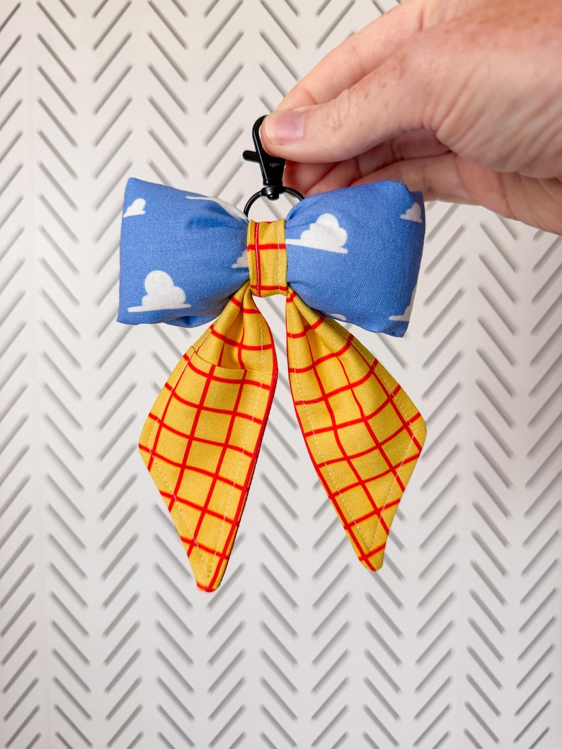 Peut inclure: Un accessoire en forme de n&oelig;ud en tissu avec un motif de nuages blancs sur fond bleu pour la partie sup&eacute;rieure et un tissu jaune &agrave; carreaux rouges pour les deux pans inf&eacute;rieurs. Le n&oelig;ud est fix&eacute; &agrave; un mousqueton pivotant en plastique noir.