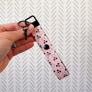 May include: Pink wristlet key fob with a black and white ghost pattern. The key fob has a black clasp and a black button. The wristlet is held by a hand.