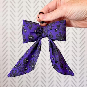 May include: A purple fabric bow keychain featuring a repeating black damask-style pattern with stylized eyes. The bow has two long, pointed tails hanging downward and is attached to a black circular metal ring for secure attachment to items.