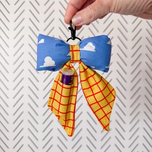 Peut inclure: Un porte-cl&eacute;s en tissu en forme de n&oelig;ud avec un motif de ciel bleu &agrave; nuages blancs sur le haut et un quadrillage jaune &agrave; lignes rouges sur les pans inf&eacute;rieurs. Un petit clip en plastique noir est fix&eacute; au sommet et un baume &agrave; l&egrave;vres est ins&eacute;r&eacute;.