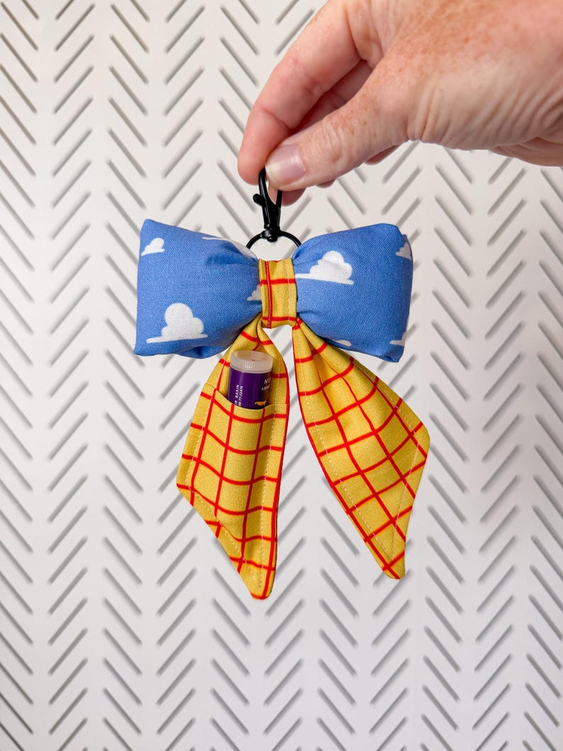 Peut inclure: Un porte-cl&eacute;s en tissu en forme de n&oelig;ud avec un motif de ciel bleu &agrave; nuages blancs sur le haut et un quadrillage jaune &agrave; lignes rouges sur les pans inf&eacute;rieurs. Un petit clip en plastique noir est fix&eacute; au sommet et un baume &agrave; l&egrave;vres est ins&eacute;r&eacute;.