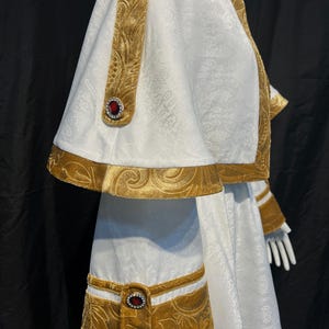 May include: White and gold ceremonial garment with intricate floral patterns. The garment features gold velvet trim and accents, including decorative bands and red jewel embellishments. The design includes a cape and long sleeves, creating a regal appearance.