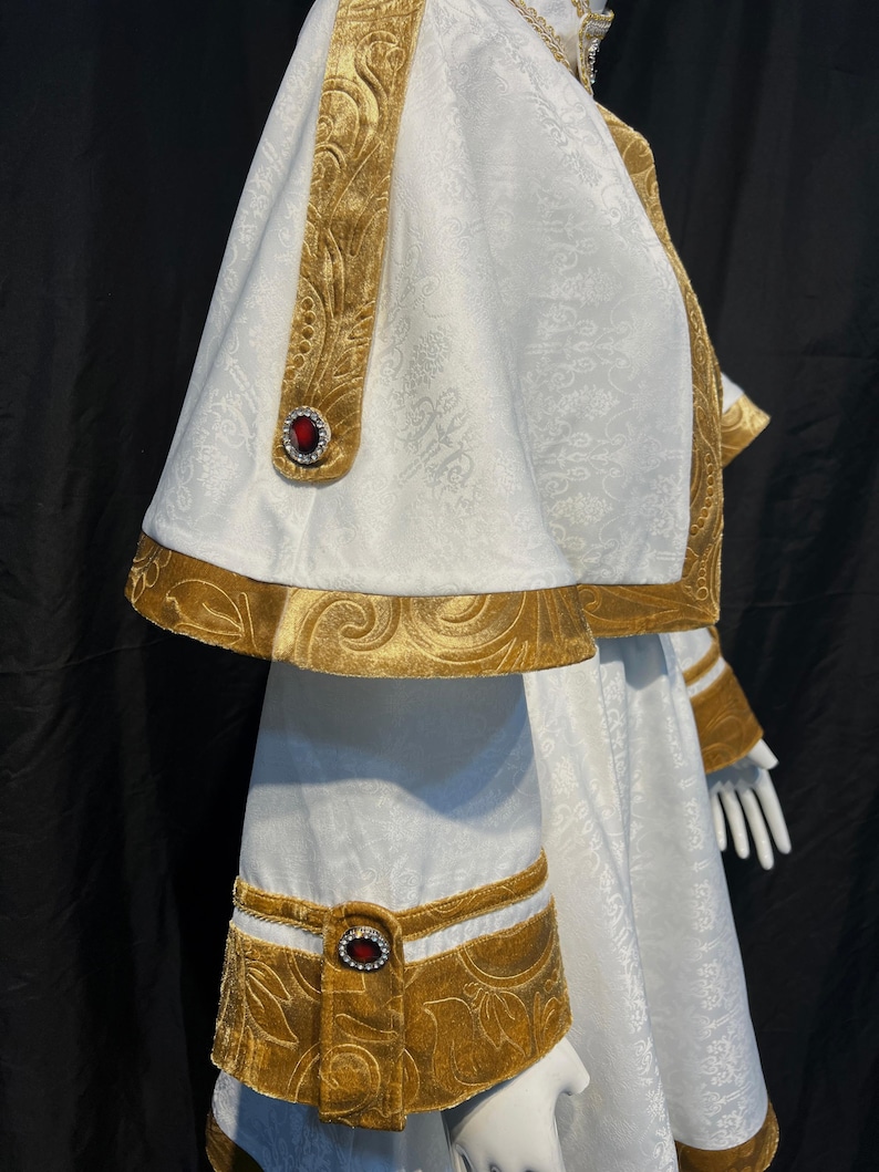 May include: White and gold ceremonial garment with intricate floral patterns. The garment features gold velvet trim and accents, including decorative bands and red jewel embellishments. The design includes a cape and long sleeves, creating a regal appearance.