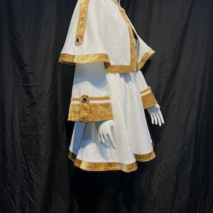 May include: A white costume with gold trim and red accents, displayed on a mannequin. The outfit includes a cape, dress, and decorative embellishments. The gold trim features an ornate pattern, and the costume is set against a black backdrop.