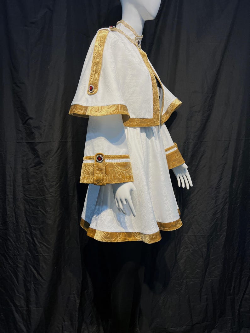 May include: A white costume with gold trim and red accents, displayed on a mannequin. The outfit includes a cape, dress, and decorative embellishments. The gold trim features an ornate pattern, and the costume is set against a black backdrop.