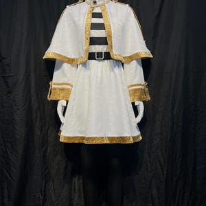 May include: A white costume with gold trim, featuring a cape and a dress. The outfit includes a black and white striped top, a black belt, and gold accents on the sleeves and cape. The costume is displayed on a mannequin.