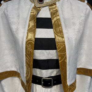 May include: A white costume with gold trim and accents. The costume features a high collar, a cape-like overlay, and a black and white striped garment underneath. A black belt with a silver buckle cinches the waist.