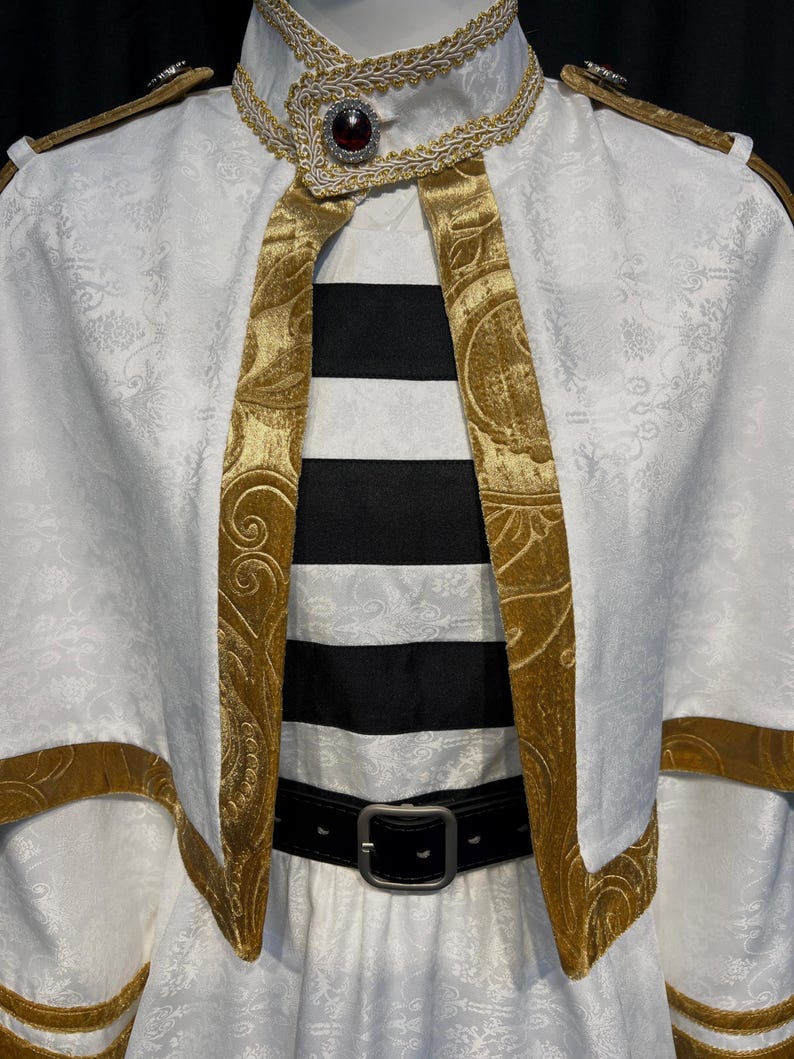 May include: A white costume with gold trim and accents. The costume features a high collar, a cape-like overlay, and a black and white striped garment underneath. A black belt with a silver buckle cinches the waist.