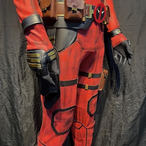 Puede incluir: Traje de Deadpool rojo y negro con cintur&oacute;n, bolsas y un s&iacute;mbolo rojo y negro en el cintur&oacute;n. El traje tiene un aspecto desgastado con detalles negros y herrajes dorados.