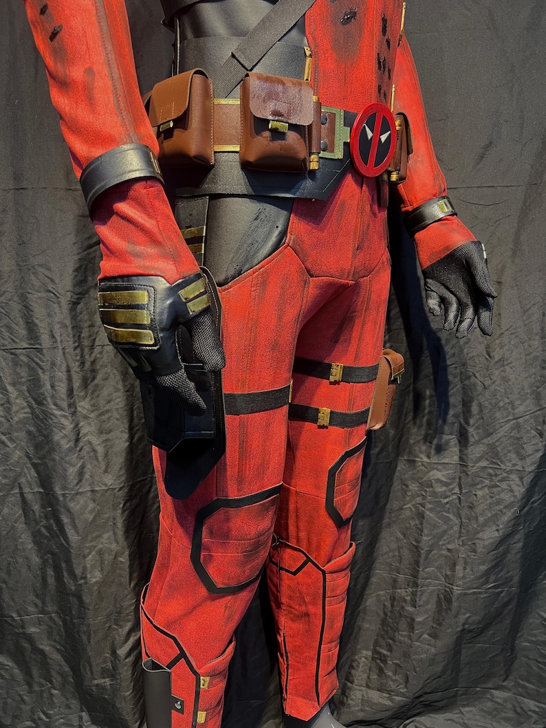 Puede incluir: Traje de Deadpool rojo y negro con cintur&oacute;n, bolsas y un s&iacute;mbolo rojo y negro en el cintur&oacute;n. El traje tiene un aspecto desgastado con detalles negros y herrajes dorados.