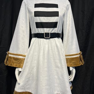 May include: White dress with long sleeves and a black belt. The dress features a black rectangular design on the bodice and gold trim on the sleeves and hem. The fabric has a subtle floral pattern.