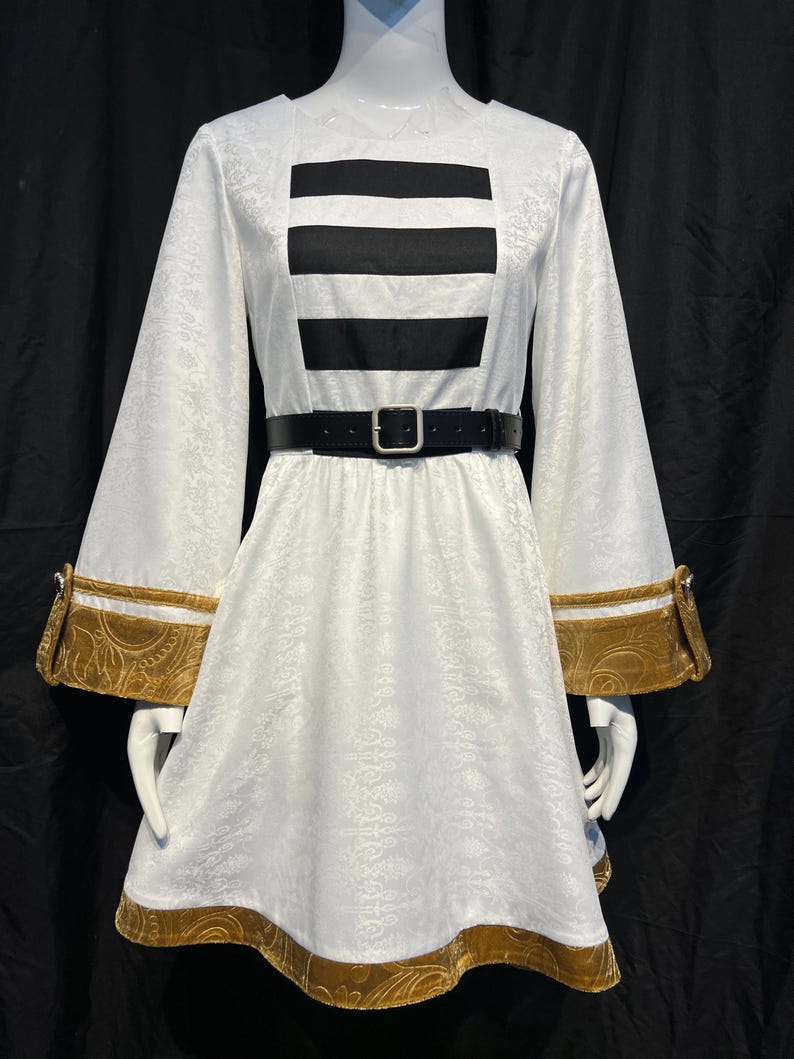 May include: White dress with long sleeves and a black belt. The dress features a black rectangular design on the bodice and gold trim on the sleeves and hem. The fabric has a subtle floral pattern.