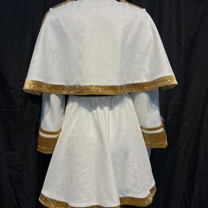 May include: A white costume with gold trim, displayed on a mannequin. The outfit includes a jacket with a cape, and a skirt. The gold trim is visible on the collar, cape, sleeves, and skirt hem. The fabric has a subtle floral pattern.