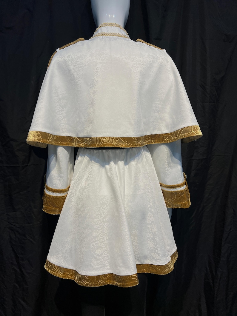May include: A white costume with gold trim, displayed on a mannequin. The outfit includes a jacket with a cape, and a skirt. The gold trim is visible on the collar, cape, sleeves, and skirt hem. The fabric has a subtle floral pattern.