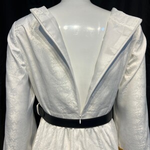 May include: A white dress with a floral pattern, long sleeves, and a black belt. The dress has gold trim on the sleeves and a zipper down the back. The dress is displayed on a mannequin.
