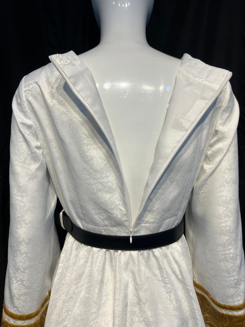 May include: A white dress with a floral pattern, long sleeves, and a black belt. The dress has gold trim on the sleeves and a zipper down the back. The dress is displayed on a mannequin.