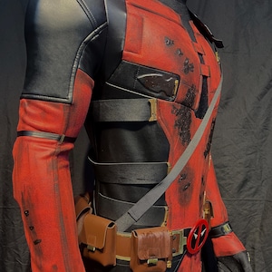 Puede incluir: Un disfraz de Deadpool rojo y negro con un cintur&oacute;n negro y bolsas marrones. El disfraz tiene un aspecto desgastado con salpicaduras de pintura marr&oacute;n y negra.
