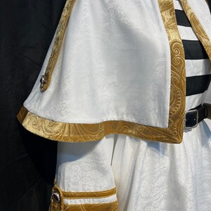 May include: White costume with gold trim and accents. The costume features a cape, decorative collar, and sleeve cuffs. The gold trim has an ornate, embossed design. The costume has a black and white striped element and a black belt.