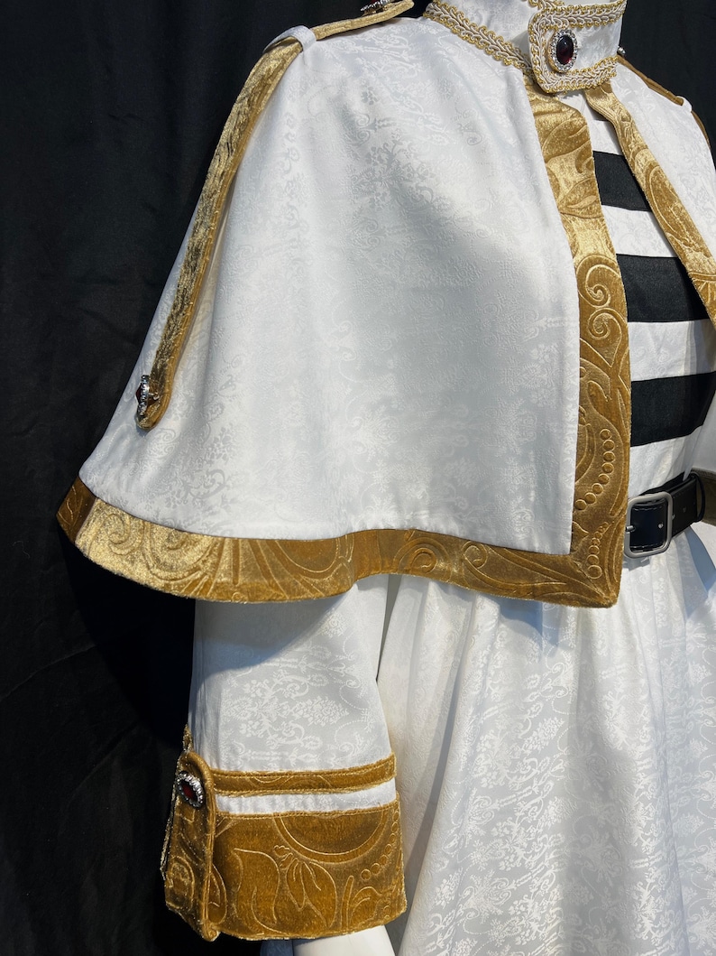 May include: White costume with gold trim and accents. The costume features a cape, decorative collar, and sleeve cuffs. The gold trim has an ornate, embossed design. The costume has a black and white striped element and a black belt.