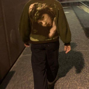 Può includere: Una persona indossa un maglione di lana verde con un ritratto di una persona sul retro. Il maglione ha maniche lunghe e un collo rotondo. La persona indossa pantaloni neri e scarpe da ginnastica bianche.