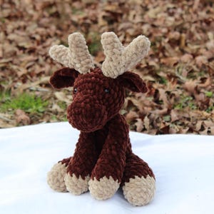 May include: A brown crocheted moose with beige antlers. The moose is sitting on a white surface.