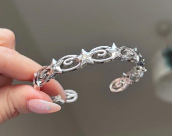 May include: A silver-toned cuff bracelet featuring a repeating pattern of stars and swirling designs. The open-ended bracelet is held up against a neutral background, showcasing its intricate details and polished finish.