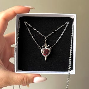 Op de afbeelding: Een zilveren ketting met een rode hart hanger met een zilveren dolk. De ketting is gelaagd met twee kettingen.