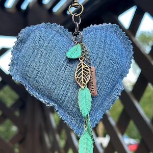 May include: A denim heart-shaped keychain with a bronze-colored clasp and chain. The chain holds three green leaf charms and a small tag that reads "breathe". The heart is made of blue denim fabric.