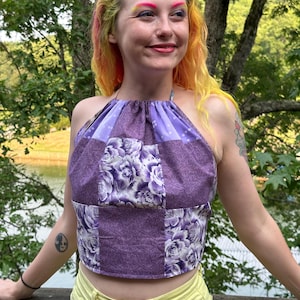 May include: A halter top featuring a patchwork design of purple fabrics with floral patterns and solid purple sections. The garment has a gathered neckline and a cropped hemline, constructed from multiple fabric panels sewn together in a square layout.