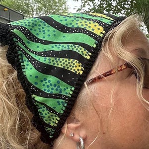 May include: A colorful headscarf with a black crocheted trim. The fabric features a pattern of green, yellow, and black stripes with polka dots. The head covering is designed to be worn on the head.
