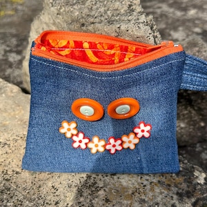 May include: A denim coin purse with an orange zipper and a handle. The purse features orange button eyes and a flower-shaped smile made of white and orange flowers. The interior lining is orange with a floral pattern.