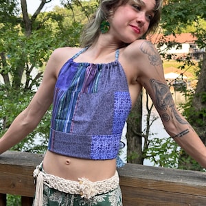 May include: A patchwork halter top featuring various shades of purple and blue fabric with paisley, floral, and striped patterns. The top has a gathered neckline and ties at the back. Below, a cream-colored crocheted belt with fringe accents.