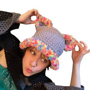 May include: A gray crocheted hat with a textured pattern and a fluffy, rainbow-colored trim. The hat has two ear-like protrusions with the same colorful trim. The hat is a unique and colorful accessory.